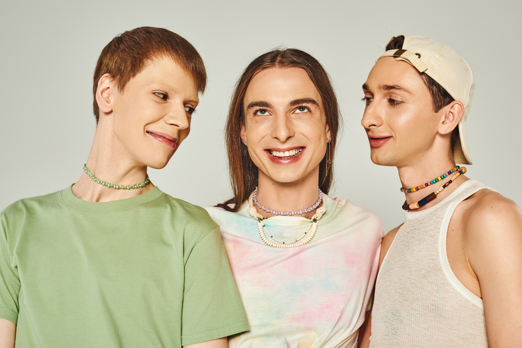 Porträt von drei lgbtq Menschen mit bunten Perlen lächelnd, während sie zusammen auf grauem Hintergrund im Studio stehen, Feier des stolzen Monats-Konzepts   - Foto, Bild