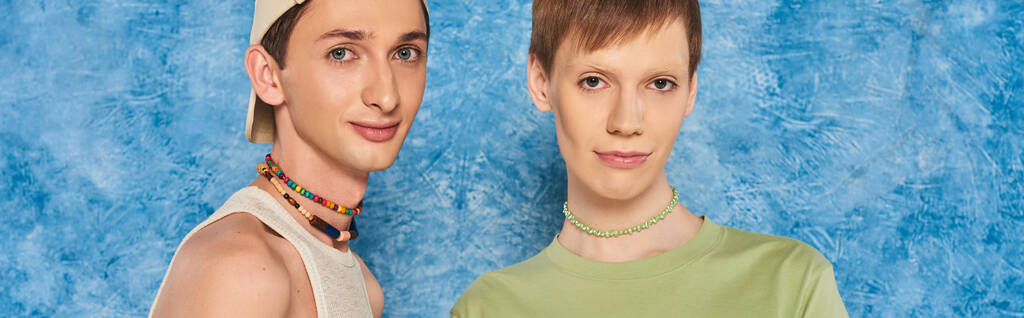 young lgbtq friends with colorful beads and casual clothes looking at camera while standing together on mottled blue background during pride month, banner  - Photo, Image