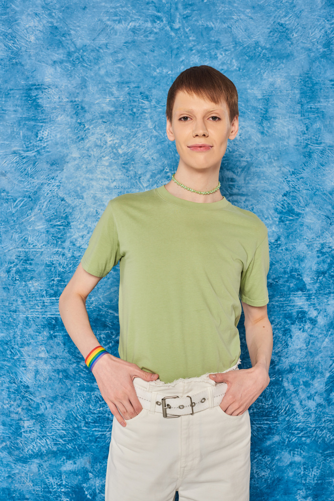 Jeune queer en vêtements décontractés tenant la main dans des poches de pantalon et regardant la caméra pendant la célébration du mois de fierté lgbt sur fond bleu moucheté - Photo, image