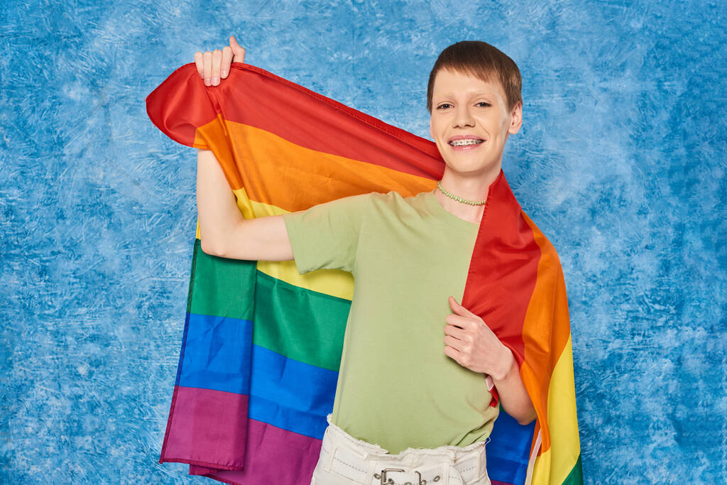 Χαρούμενος gay άντρας με casual ρούχα χαμογελώντας και κρατώντας τη σημαία Igbt και κοιτάζοντας την κάμερα κατά τη διάρκεια της γιορτής μήνα υπερηφάνειας σε στίγματα μπλε φόντο - Φωτογραφία, εικόνα