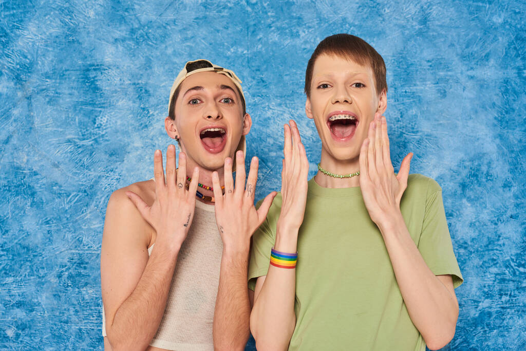 Σοκαρισμένοι ομοφυλόφιλοι φίλοι σε casual ρούχα ανοίγοντας το στόμα και κοιτάζοντας την κάμερα κατά τη διάρκεια Igbt γιορτή μήνα υπερηφάνειας για στίγματα μπλε φόντο - Φωτογραφία, εικόνα