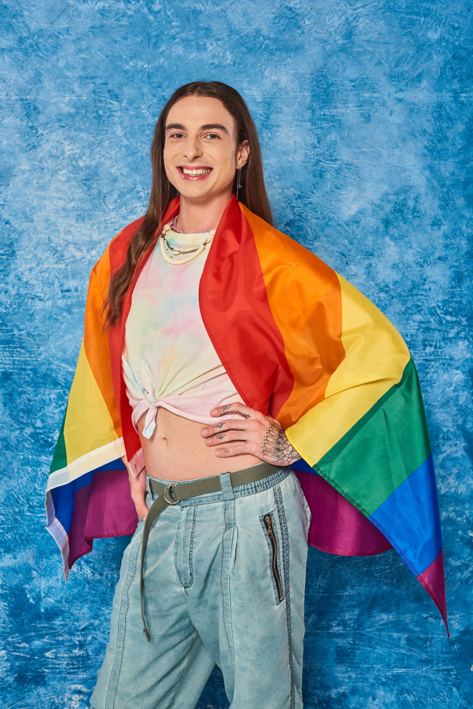 Szczęśliwy długowłosy i wytatuowany gej człowiek w casual ubrania z flagą lgbt pozowanie i patrząc w kamerę podczas świętowania dumy miesiąc na cętkowane niebieskie tło - Zdjęcie, obraz