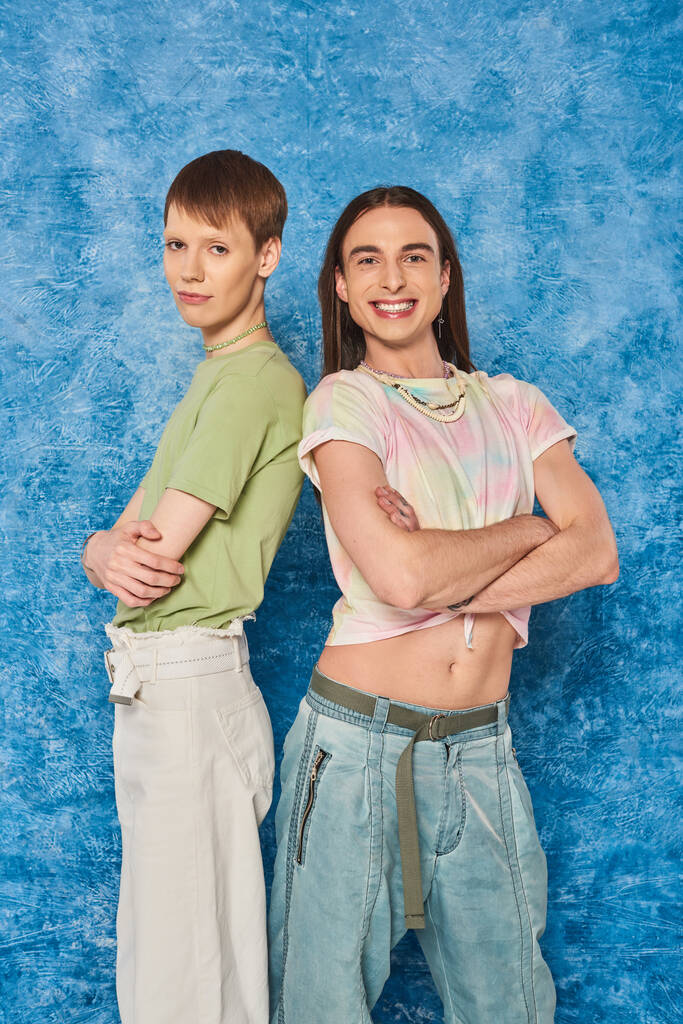 Lächelnder langhaariger schwuler Mann, der die Arme kreuzt, während er neben einem homosexuellen Freund steht, während der lgbt Pride Monatsfeier auf texturiertem blauem Hintergrund - Foto, Bild