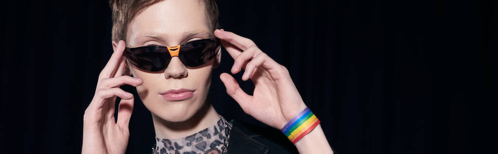 Portrait of trendy nonbinary person with lgbt flag on bracelet touching sunglasses during pride community month celebration isolated on black, banner  - Photo, Image