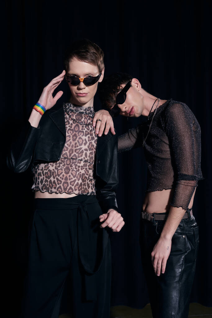 Μοντέρνοι και νεαροί γκέι φίλοι με γυαλιά ηλίου και ρούχα για πάρτι ποζάρουν και κοιτάζουν την κάμερα κατά τη διάρκεια της γιορτής του μήνα υπερηφάνειας του Igbt απομονωμένοι σε μαύρο - Φωτογραφία, εικόνα