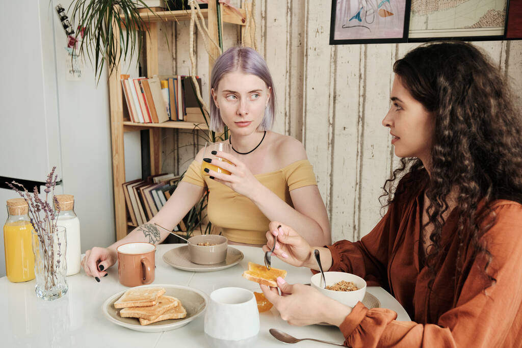Όμορφες λεσβίες που κάθονται σε στρογγυλό τραπέζι και τρώνε προπόσεις ενώ τρώνε πρωινό στο σπίτι - Φωτογραφία, εικόνα