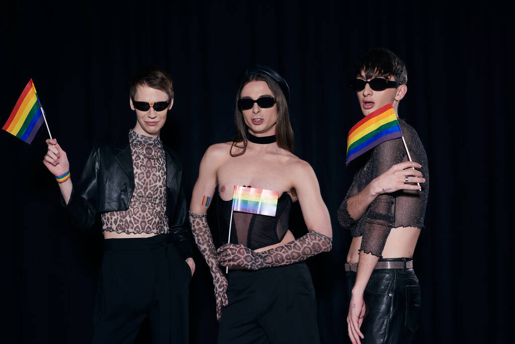 Молодые и стильные друзья-гомосексуалисты в солнцезащитных очках и костюмах для вечеринок с флагами lgbt во время празднования месяца гордости, изолированного на черном  - Фото, изображение