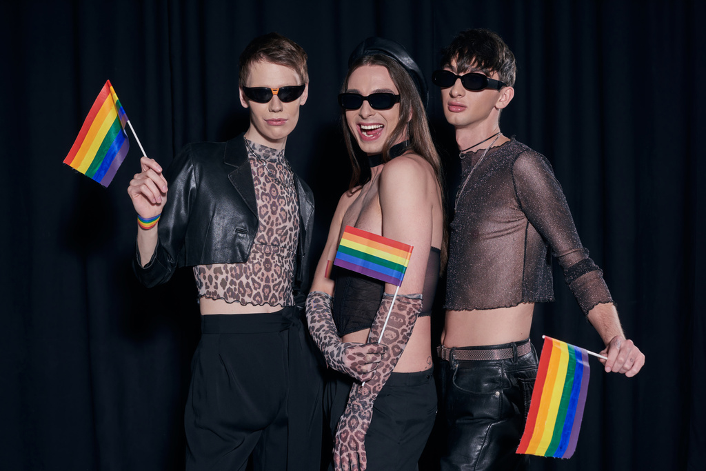 Μοντέρνοι και θετικοί ομοφυλόφιλοι φίλοι με ρούχα κόμμα και γυαλιά ηλίου ποζάρουν με σημαίες Igbtq κατά τη διάρκεια εορτασμού μήνα υπερηφάνειας σε μαύρο φόντο  - Φωτογραφία, εικόνα