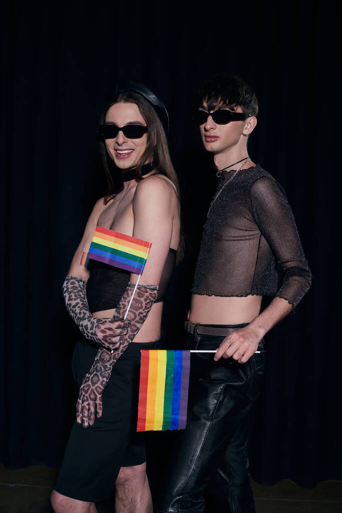 Modne homoseksualne przyjaciółki w strojach imprezowych i okularach przeciwsłonecznych z tęczowymi flagami i pozujące podczas obchodów miesiąca flagi lgbt izolowane na czarno  - Zdjęcie, obraz