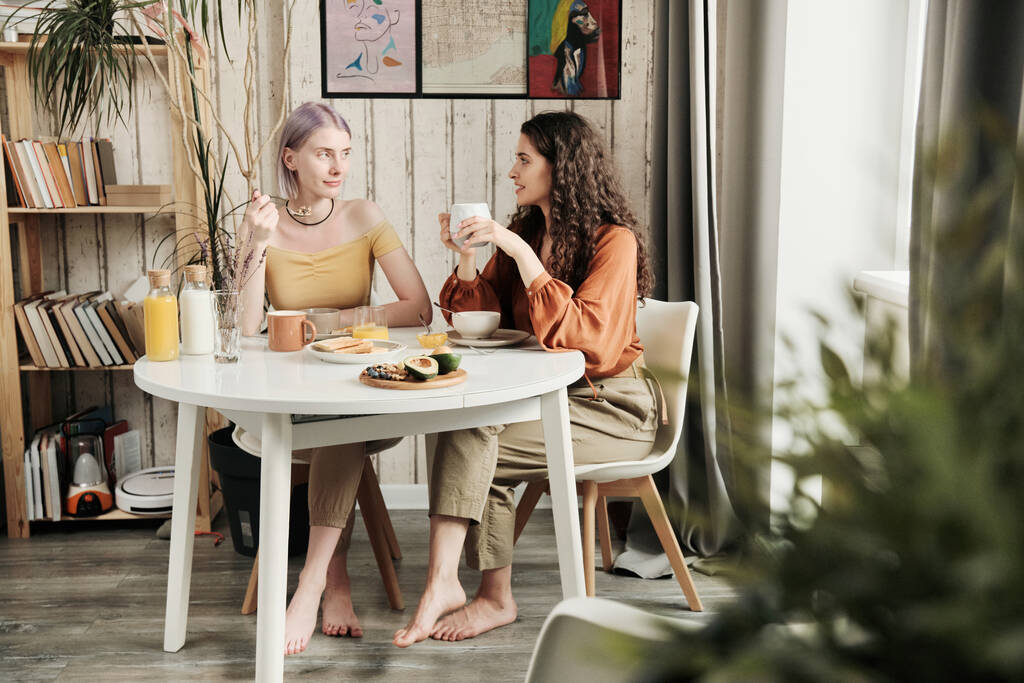 Aantrekkelijke jonge lesbiennes zitten aan tafel en drinken koffie tijdens het ontbijt thuis - Foto, afbeelding
