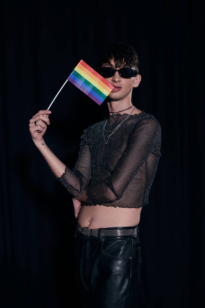 modny homoseksualny młody mężczyzna w okulary przeciwsłoneczne i błyszczący top gryzienie flaga lgbt i pozowanie podczas dumy społeczności miesiąc uroczystości odizolowany na czarny  - Zdjęcie, obraz