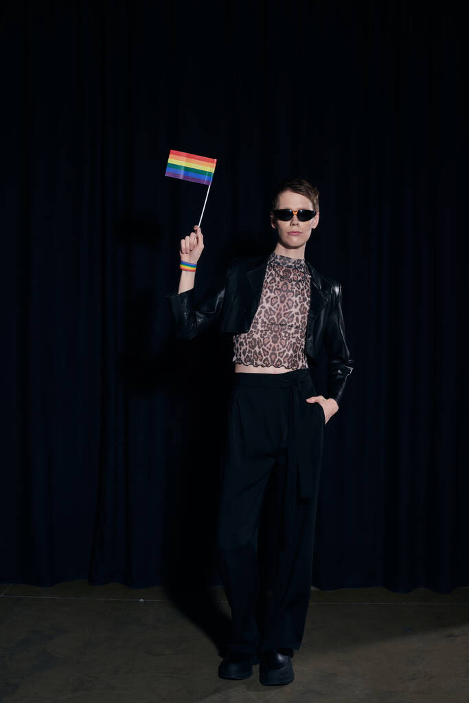 Full length of stylish nonbinary person in party outfit and sunglasses holding rainbow flag and posing during lgbt pride month celebration on black background  - Photo, Image