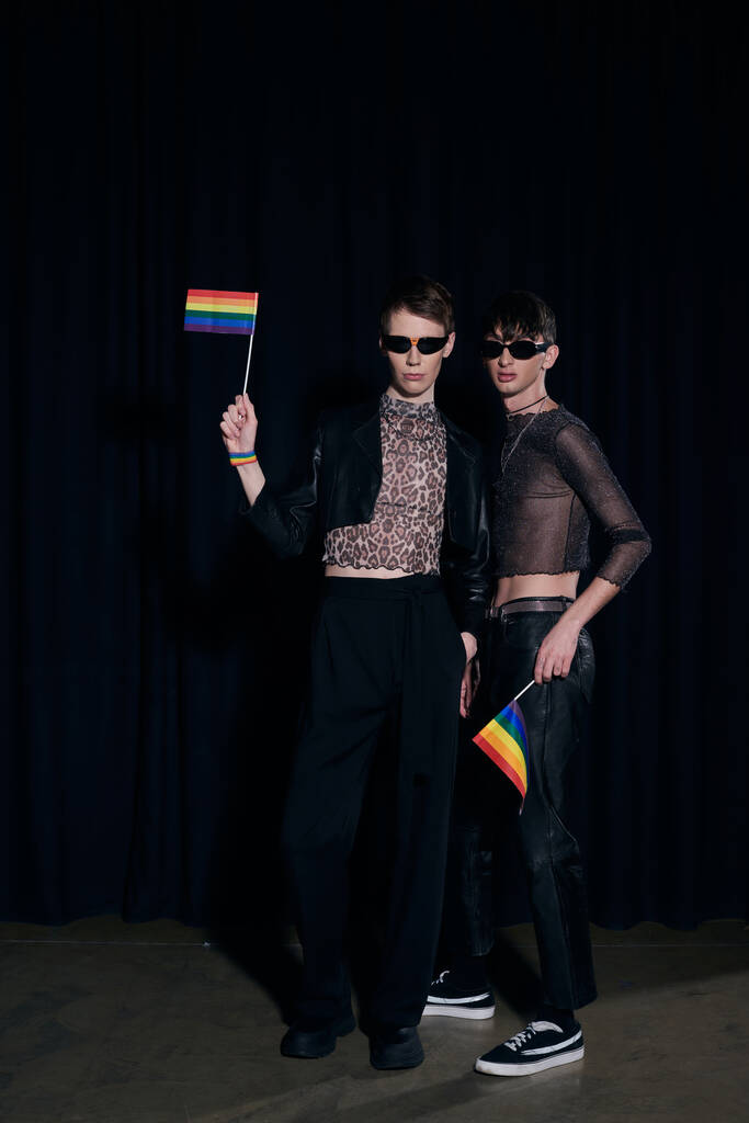 Full length of fashionable gay friends in sunglasses and party outfits holding rainbow lgbt flags during pride community month celebration on black background  - Photo, Image