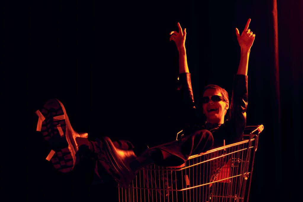 Aufgeregte nichtbinäre Person mit Sonnenbrille und stylischem Outfit, die mit den Fingern zeigt, während sie im Einkaufswagen sitzt, während lgbt Stolz Monatsfeier auf schwarzem Hintergrund mit rotem Licht  - Foto, Bild
