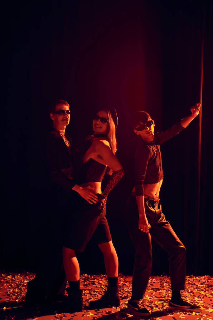 In voller Länge lächelnde und modische homosexuelle Menschen mit Sonnenbrille posieren auf Konfetti während der Party und lgbt Stolz Monatsfeier auf schwarzem Hintergrund mit rotem Licht  - Foto, Bild