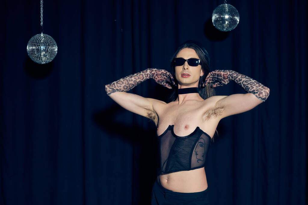 Модный гей в сексуальном корсете и солнцезащитных очках позирует возле дискотечных мячей во время вечеринки и празднования месяца гордости на черном фоне  - Фото, изображение