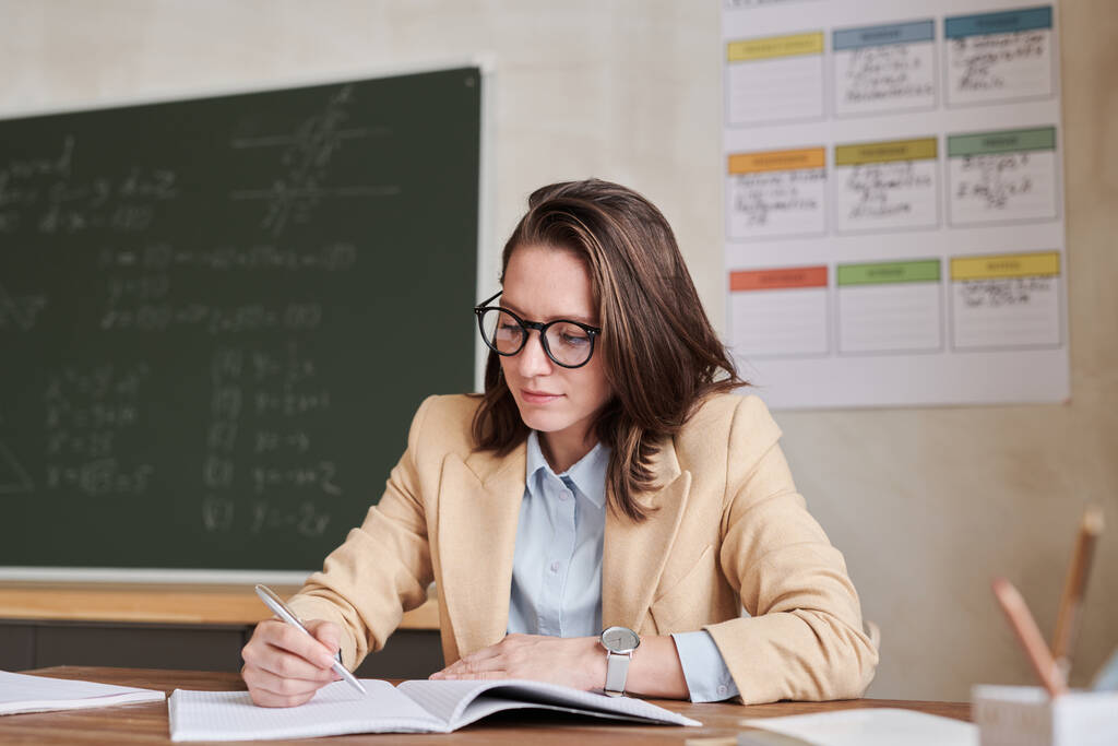 Warm gefärbtes Porträt einer jungen Lehrerin, die im Klassenzimmer am Schreibtisch sitzt und schreibt, Kopierraum - Foto, Bild
