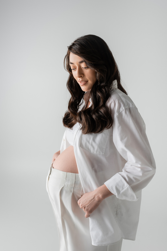 очаровательная будущая мама в белой модной одежде, такой как белая рубашка, кукурузный топ и брюки, улыбающиеся изолированно на сером фоне, стильная концепция материнства, беременность  - Фото, изображение