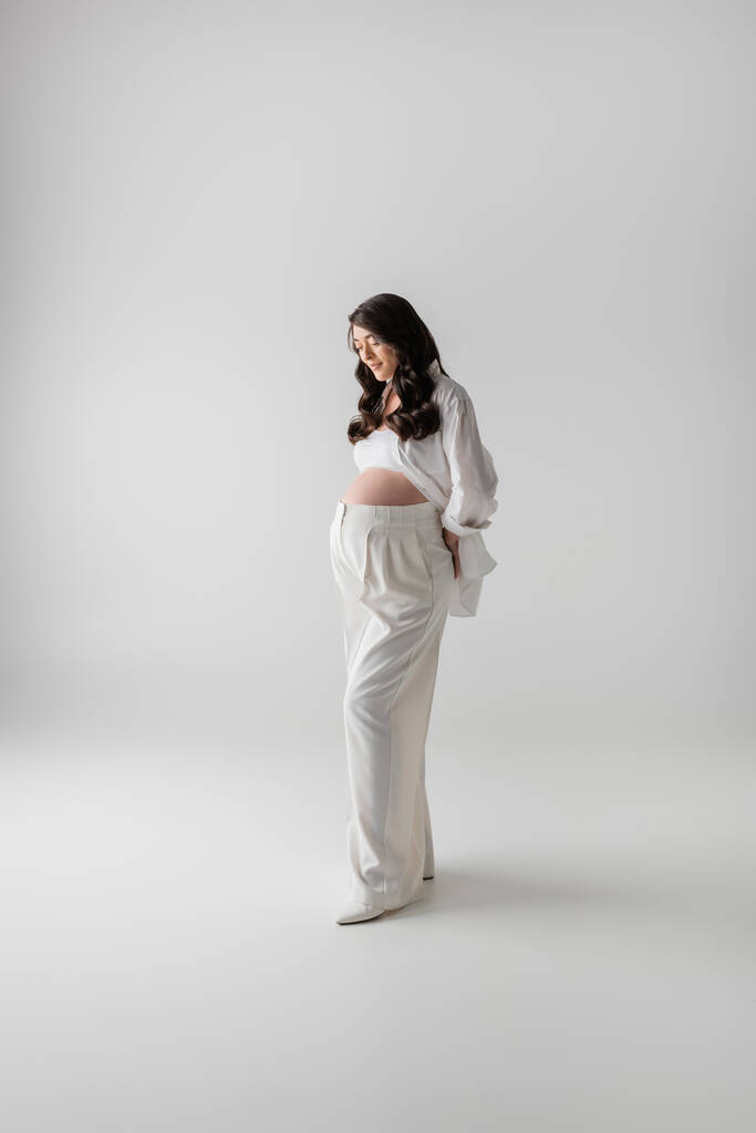 pełna długość brunetka i atrakcyjna ciężarna modelka w białych stylowych spodniach i koszuli trzymając się za plecy stojąc na szarym tle, modna koncepcja macierzyństwa - Zdjęcie, obraz