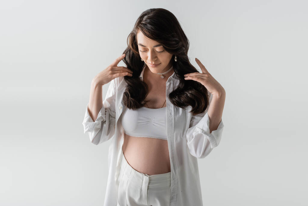joyful pregnant woman in white crop top, stylish shirt and pants touching wavy brunette hair and looking down isolated on grey background, maternity fashion concept - Photo, Image