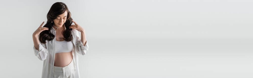 mulher grávida elegante em camisa branca da moda, top de cultura e calças tocando cabelos ondulados morena e posando isolado em fundo cinza, conceito de maternidade na moda, banner - Foto, Imagem