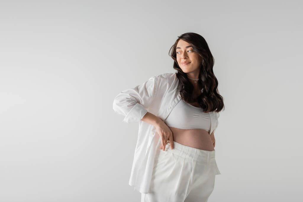 studio shot of pleased future mother in white stylish shirt holding white pants, smiling and looking away isolated on grey background, maternity fashion concept - Photo, Image