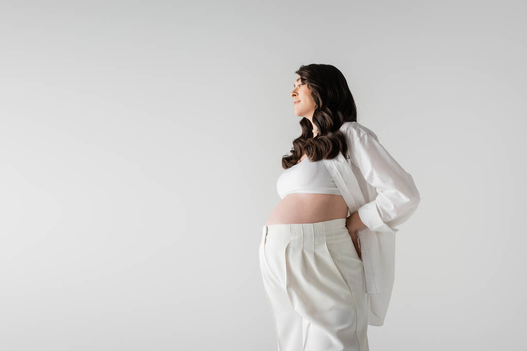 side view of smiling pregnant woman with wavy brunette hair, wearing white stylish maternity clothes, posing with hand behind back isolated on grey background, fashionable maternity concept - Photo, Image