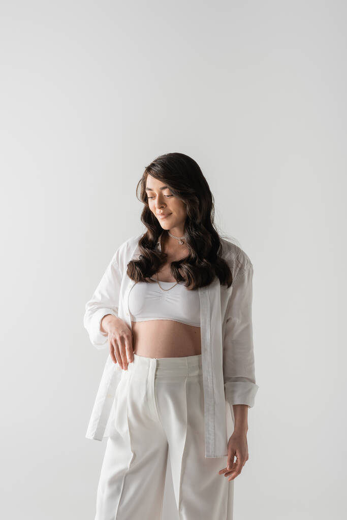 zufriedene schwangere Frau mit welligem brünetten Haar, weißes bauchfreies Oberteil, Hemd und Hose, stehend und lächelnd isoliert auf grauem Hintergrund, Umstandsmodekonzept - Foto, Bild