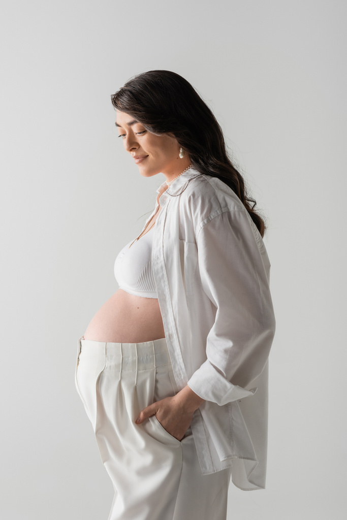 Seitenansicht der glücklichen werdenden Mutter in weißem Crop Top, Hemd und Hose posieren mit der Hand in der Tasche, isoliert auf grauem Hintergrund, modisches Umstandskonzept, schwangere Frau  - Foto, Bild