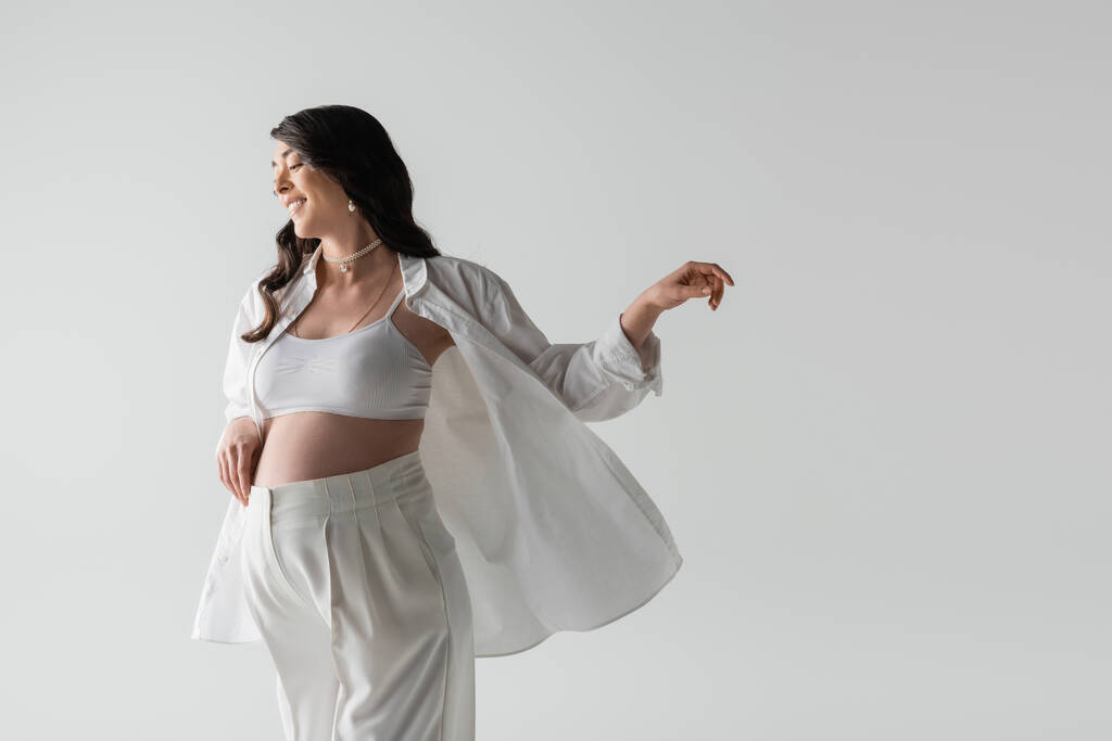 positive brunette pregnant woman in shirt, crop top and pants posing with outstretched hand isolated on grey background, fashionable pregnancy concept - Photo, Image