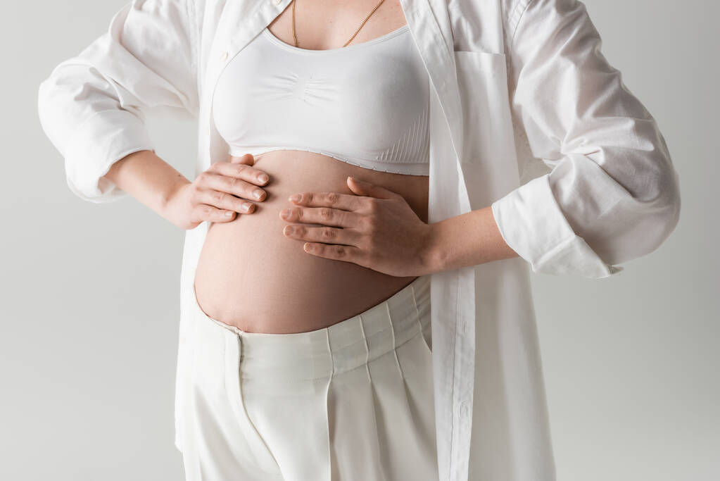 partial view of fashionable expecting mother in crop top, shirt and pants posing with hands on tummy isolated on grey background, maternity style concept, pregnant woman  - Photo, Image