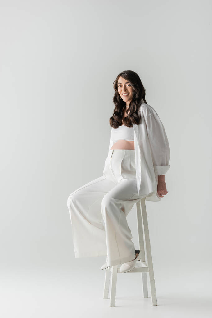 full length of pleased expected mother in white fashion pants, crop top and shirt sitting on stool and looking at camera on grey background, έννοια μόδας μητρότητας, έγκυος γυναίκα  - Φωτογραφία, εικόνα