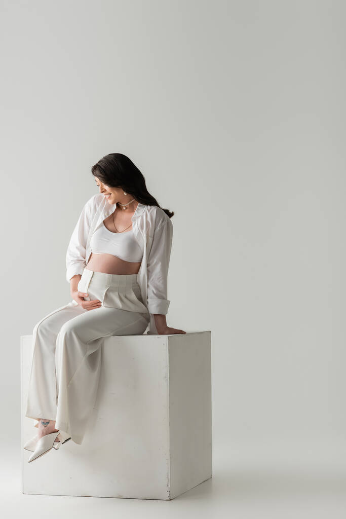 Stüdyoda, gri arka planda beyaz bir kübün üzerinde otururken şık pantolonlar içinde gülümseyen anne ve gömleğin göbeğine dokunması, hamilelik modası konsepti, hamile kadın.  - Fotoğraf, Görsel