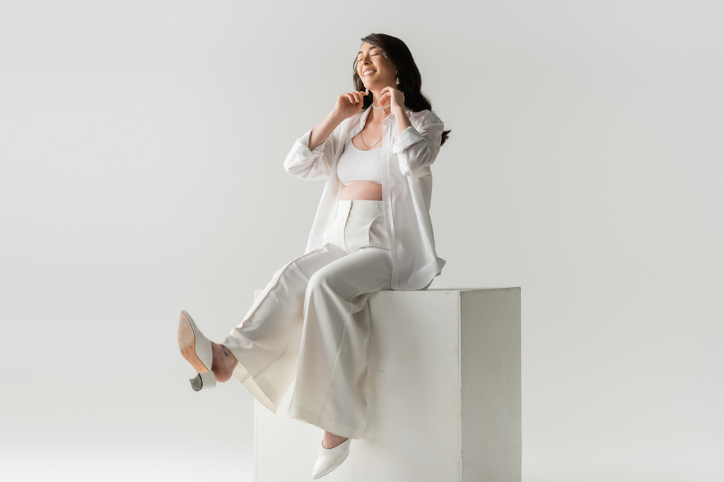 full length of joyful expecting mother in trendy pants, shirt and cropped top sitting on white cube and smiling with closed eyes on grey background, maternity fashion concept - Photo, Image
