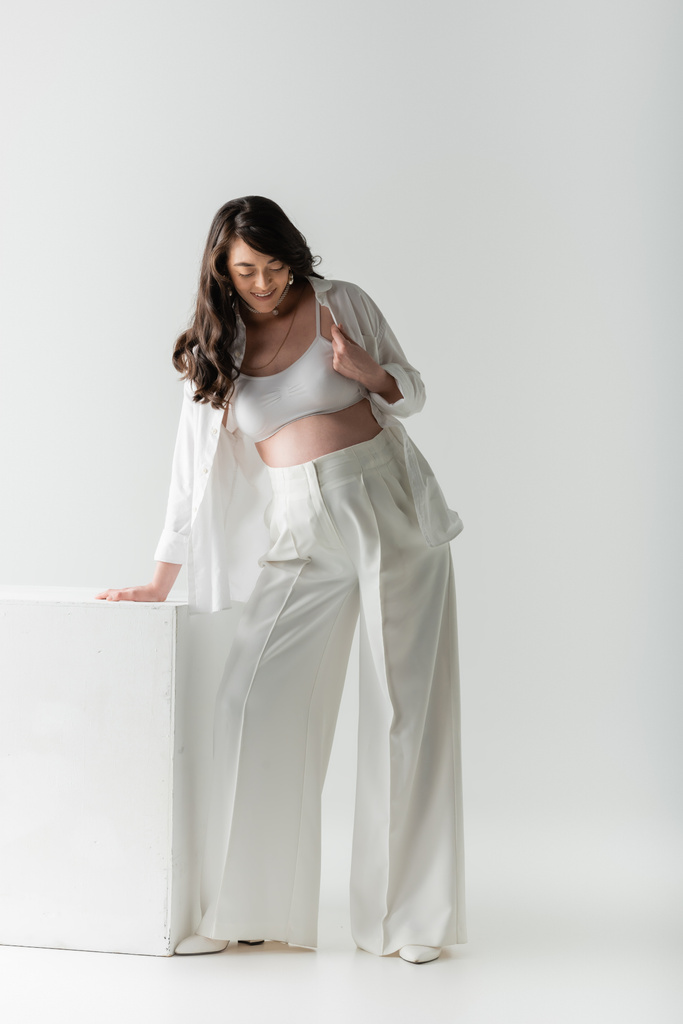 full length of pleased pregnant woman with wavy brunette hair, wearing white pants, crop top and shirt, standing near cube on grey background, maternity fashion concept - Photo, Image
