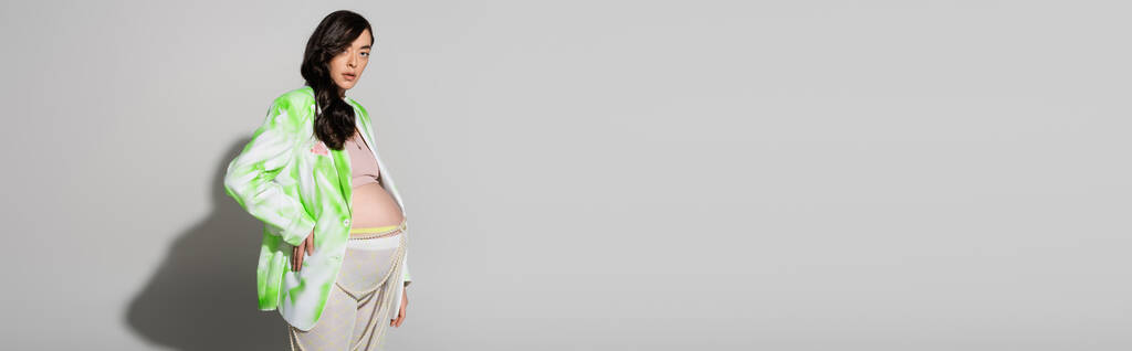 élégante future maman avec cheveux bruns ondulés, veste verte et blanche, leggings et perles ceinture debout avec la main sur la hanche sur fond gris, concept de mode maternité, bannière, femme enceinte  - Photo, image