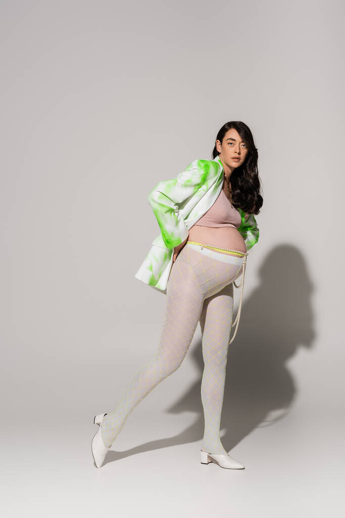 Uzun boylu esmer, külotlu çorap giymiş, yeşil ve beyaz ceketli hamile bir kadın gri arka planda poz veriyor, annelik modası, beklenti...  - Fotoğraf, Görsel