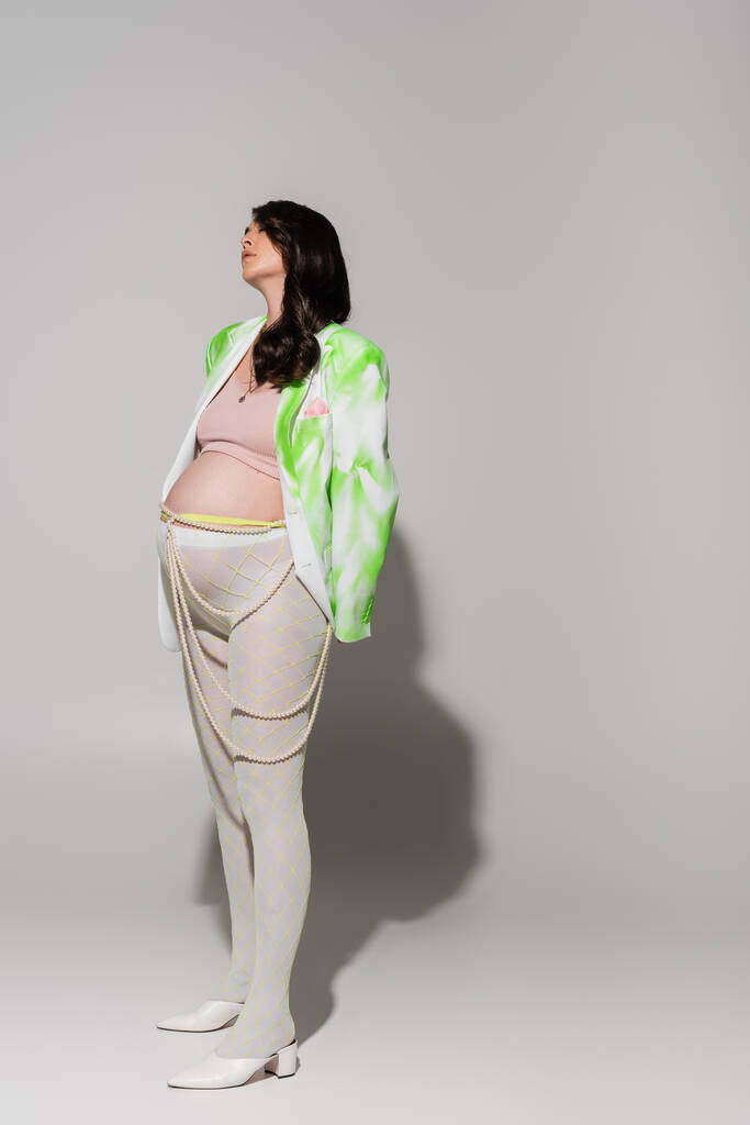 longitud completa de la mujer embarazada de moda en medias, top de la cosecha, chaqueta verde y blanco y cinturón de perlas de pie sobre fondo gris, concepto de moda de maternidad, expectativa  - Foto, imagen