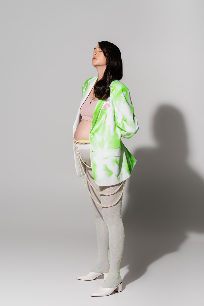 full length of pregnant woman with closed eyes standing in green and white jacket, crop top, beads belt and leggings on grey background, maternity style concept, expectation - Photo, Image
