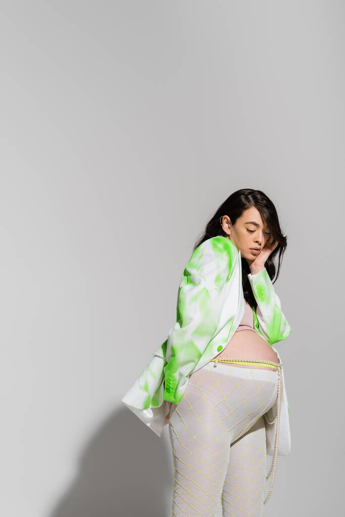 charming expecting mother in green and white blazer, crop top, leggings and beads belt touching face and posing with hand behind back on grey background, expectation, maternity fashion concept - Photo, Image