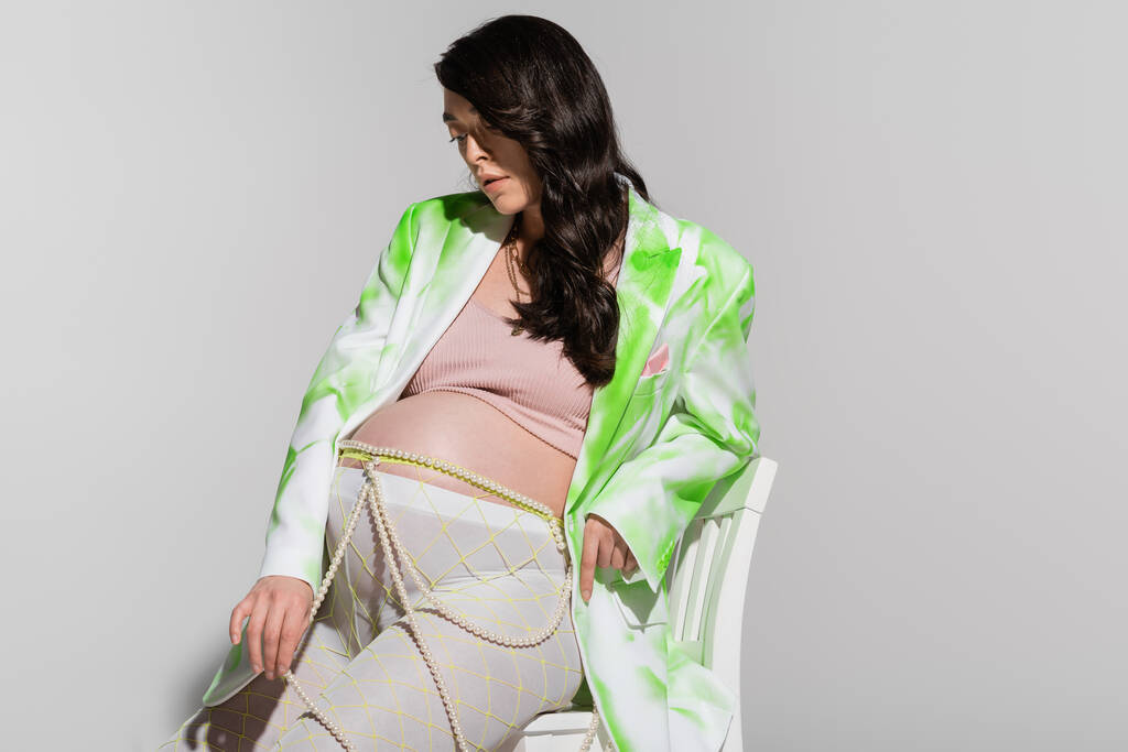 encantadora morena mamá-a-ser en blazer verde y blanco, top de la cosecha, leggings y abalorios cinturón sentado en la silla sobre fondo gris, concepto de maternidad de moda, expectativa - Foto, imagen