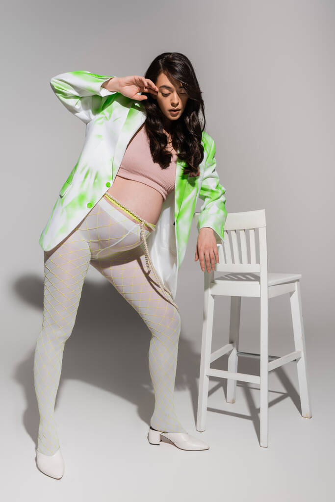volledige lengte van charmante zwangere vrouw in groene en witte blazer, gewas top, kralen riem en leggings poseren in de buurt stoel op grijze achtergrond, modieuze zwangerschap concept, verwachting - Foto, afbeelding