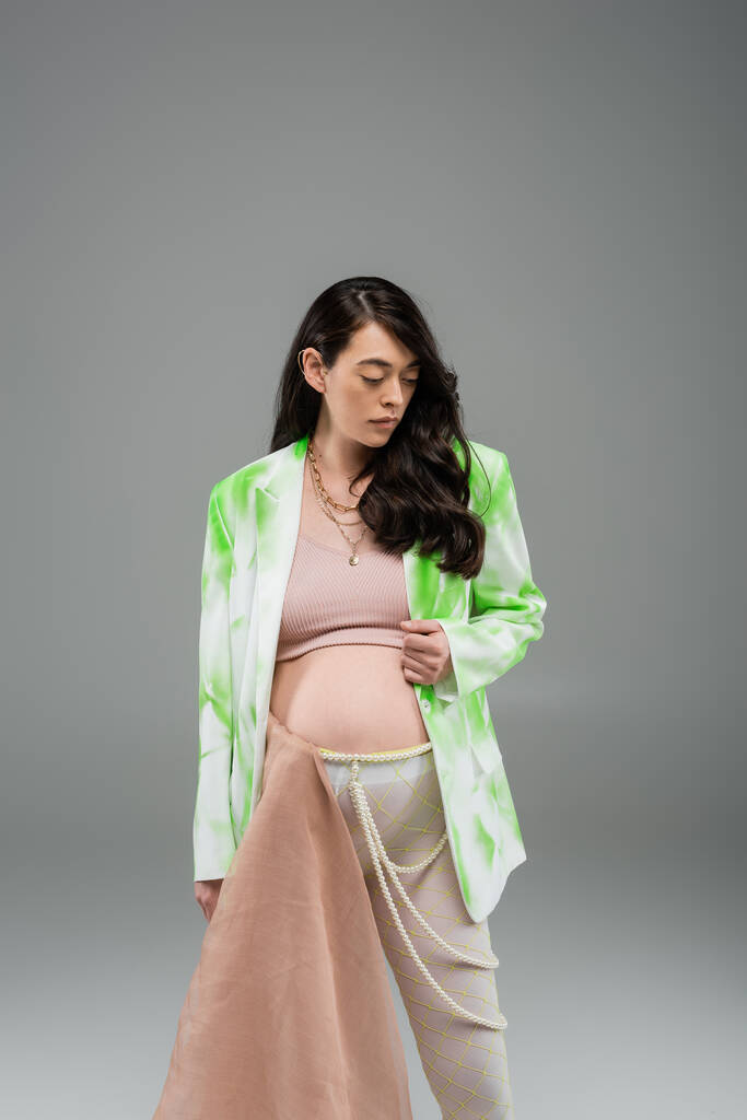 morena mujer embarazada en blazer verde y blanco, top de la cosecha, cinturón de cuentas y leggings con tela de gasa beige sobre fondo gris, concepto de moda de maternidad - Foto, imagen