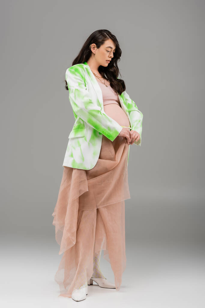 妊娠中の女性の完全な長さの波状ブルネットの髪のポーズで緑と白のブレザー、作物の上とベージュのシフォン布でグレーの背景、マタニティファッションコンセプト、期待 - 写真・画像