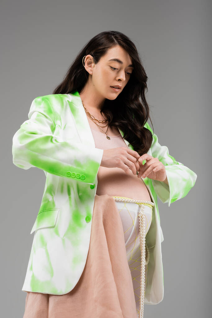 madre embarazada en chaqueta verde y blanca, top de la cosecha, leggins con cinturón de cuentas y tela de gasa posando con las manos cerca de la barriga aislada sobre fondo gris, concepto de maternidad de moda - Foto, imagen