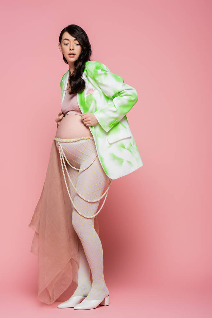 妊娠中の女性の完全な長さとともに波状ブルネットの髪ポージングでファッショナブルなブレザー、ビーズベルトとレギンスとともにベージュシフォン布でピンクの背景、マタニティファッションコンセプト - 写真・画像