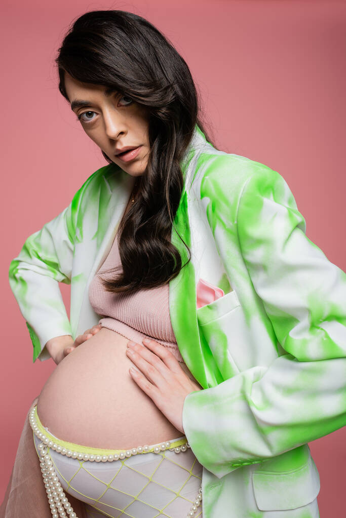 kobieta w ciąży z falowanymi brunetkami, ubrana w zieloną i białą marynarkę, top i pasek z koralikami, obejmujący brzuch i patrząc na aparat odizolowany od różowej, stylowej koncepcji macierzyństwa - Zdjęcie, obraz