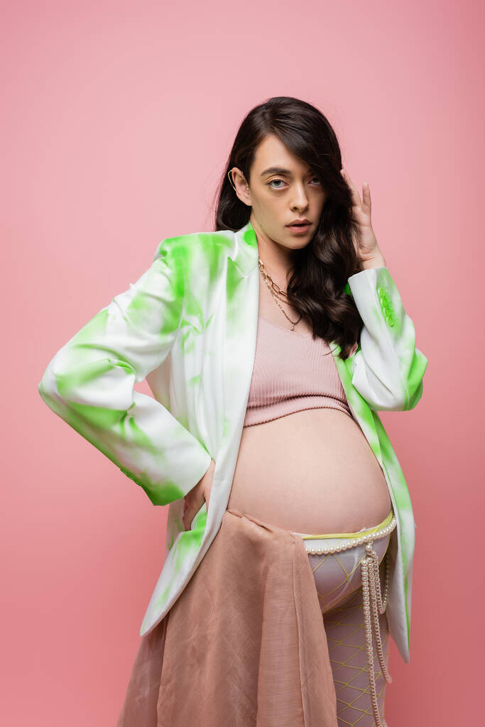 atraente futura mãe em blazer verde e branco, top de cultura e leggings com cinto de contas e pano de chiffon olhando para a câmera isolada no fundo rosa, conceito de gravidez elegante - Foto, Imagem
