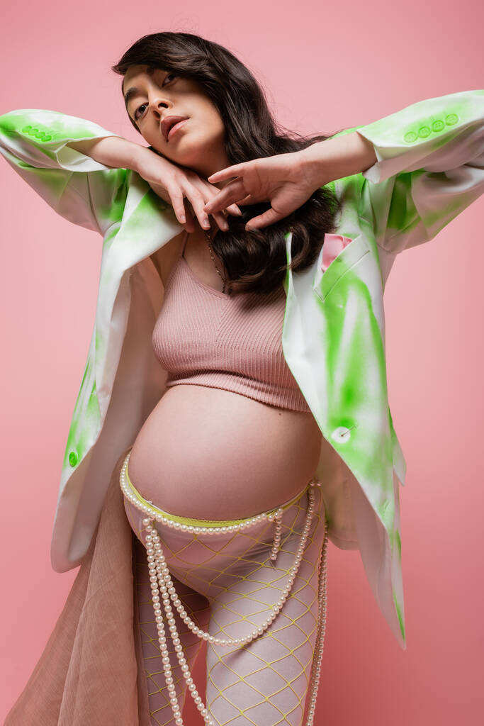 brunette pregnant woman in green and white blazer, crop top, leggings with beads belt and chiffon cloth holding hands under chin and looking at camera isolated on pink, maternity fashion concept - Photo, Image