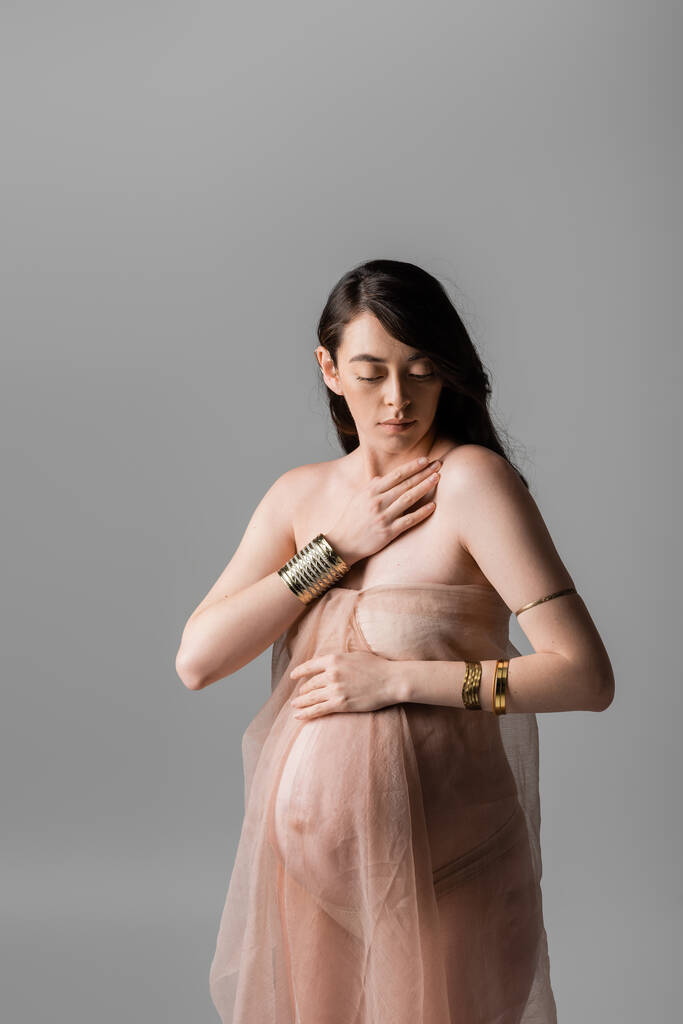 barna terhes nő arany kiegészítők megérintése mellkas közben pózol gyengéd sifon draping elszigetelt szürke háttér, anyaság stílus koncepció, érzékiség - Fotó, kép
